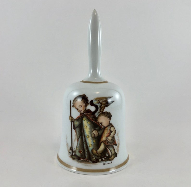 May include: White porcelain bell with a gold trim. The bell features a painting of an angel with wings and a child. The angel is holding a staff and the child is holding the angel's hand. The painting is signed "Hummel".