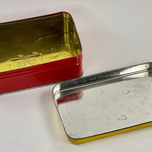 May include: A red and gold metal tin with the text "K-2013 MADE IN THAILAND" printed on the bottom. The tin is open and the inside is empty.