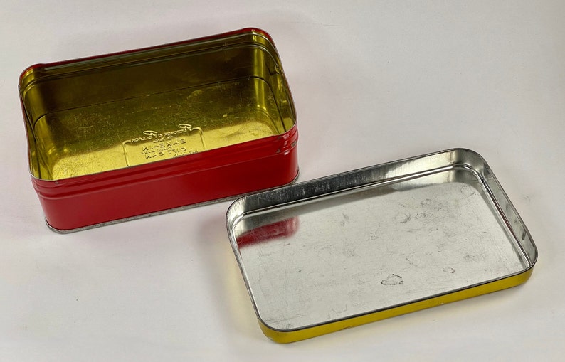 May include: A red and gold metal tin with the text "K-2013 MADE IN THAILAND" printed on the bottom. The tin is open and the inside is empty.