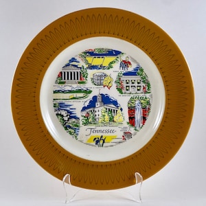 May include: A yellow and white ceramic plate with a decorative border. The center of the plate features a colorful illustration of landmarks in Tennessee, including the state capitol building, the Hermitage, and the Great Smoky Mountains. The text "Tennessee" is printed in the center of the plate.