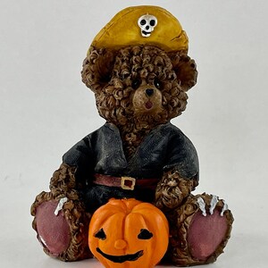 May include: A brown teddy bear wearing a black costume and a yellow pirate hat with a skull and crossbones design. The bear is holding an orange jack-o'-lantern.