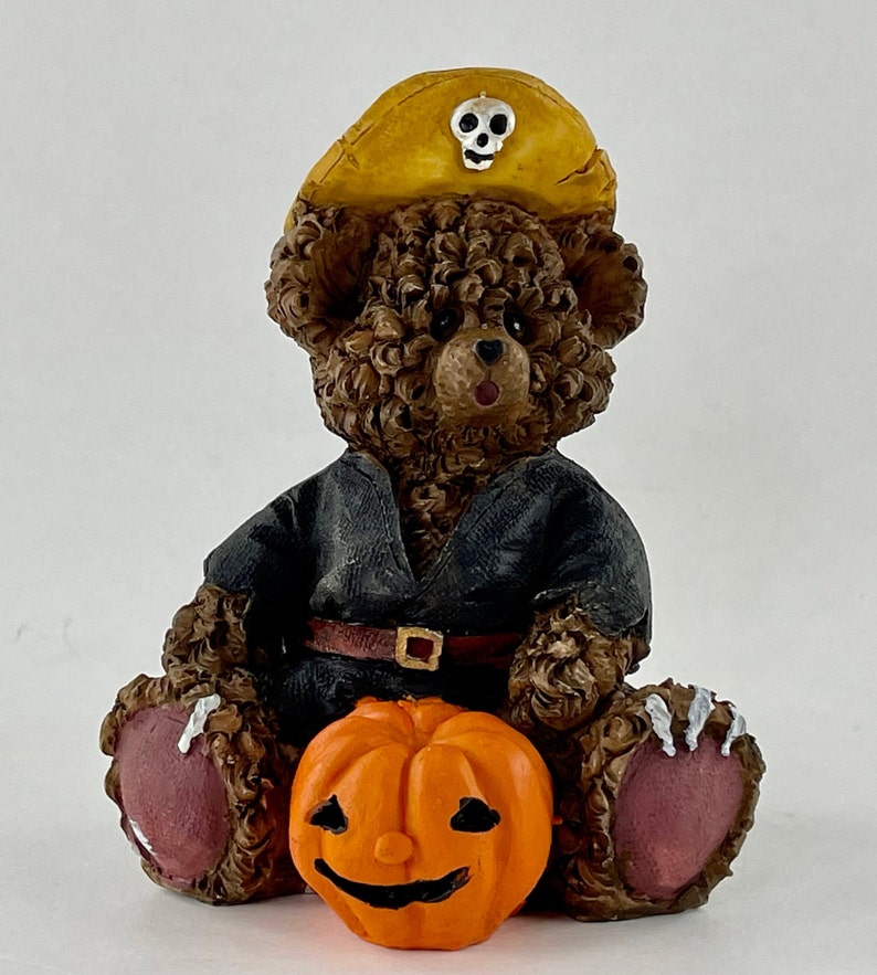 May include: A brown teddy bear wearing a black costume and a yellow pirate hat with a skull and crossbones design. The bear is holding an orange jack-o'-lantern.