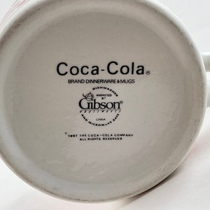 Op de afbeelding: Witte keramische Coca-Cola mok met een handvat. De onderkant van de mok toont het Coca-Cola logo, merkinformatie en het Gibson Housewares logo. De mok is vaatwasser- en magnetronbestendig.