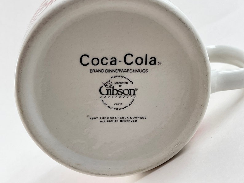 Op de afbeelding: Witte keramische Coca-Cola mok met een handvat. De onderkant van de mok toont het Coca-Cola logo, merkinformatie en het Gibson Housewares logo. De mok is vaatwasser- en magnetronbestendig.