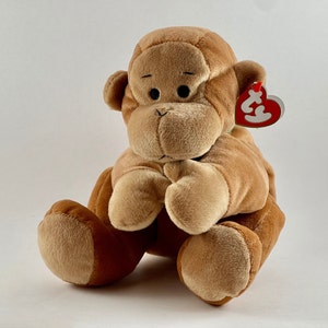 May include: A brown plush monkey toy with a red beanie baby tag. The monkey is sitting with its hands clasped together in front of it.