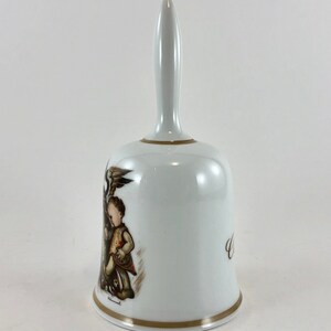 May include: White porcelain bell with a brown and gold design featuring a child holding a bird. The bell has a gold trim and the inscription "Welcome" on the side.