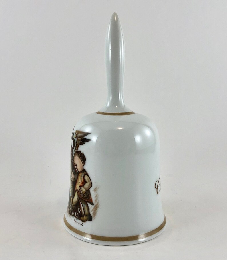 May include: White porcelain bell with a brown and gold design featuring a child holding a bird. The bell has a gold trim and the inscription "Welcome" on the side.