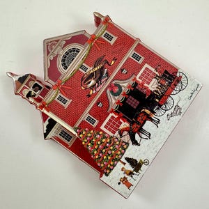 May include: A red and white wooden Christmas ornament shaped like a firehouse with a Christmas tree in front. The firehouse has a bell tower, a sign that says "Protection", and a wreath on the door. There are horses and a carriage in front of the firehouse.