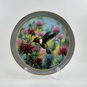 May include: A decorative plate featuring a hummingbird in flight, hovering near pink flowers. The plate has a gray border with a gold trim. The background includes blue and yellow hues, creating a vibrant scene. The plate is displayed on a clear stand.