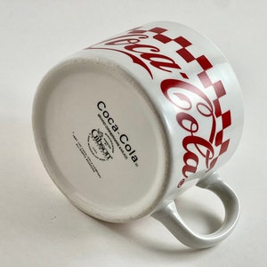Op de afbeelding: Witte keramische Coca-Cola mok met een rood geblokt patroon en Coca-Cola logo. De mok heeft een gebogen oor en het Coca-Cola logo herhaalt zich rond de rand. De onderkant van de mok heeft de merknaam Coca-Cola en het Gibson logo.