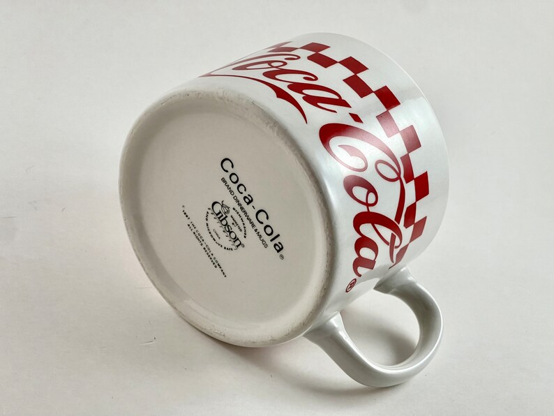 Op de afbeelding: Witte keramische Coca-Cola mok met een rood geblokt patroon en Coca-Cola logo. De mok heeft een gebogen oor en het Coca-Cola logo herhaalt zich rond de rand. De onderkant van de mok heeft de merknaam Coca-Cola en het Gibson logo.