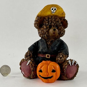 May include: A brown teddy bear wearing a black costume and a yellow pirate hat with a skull and crossbones design. The bear is holding a small orange pumpkin with a carved jack-o'-lantern face.