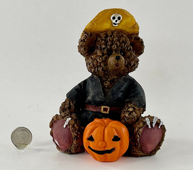 May include: A brown teddy bear wearing a black costume and a yellow pirate hat with a skull and crossbones design. The bear is holding a small orange pumpkin with a carved jack-o'-lantern face.