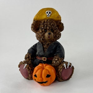 May include: A brown teddy bear wearing a black pirate costume and a yellow pirate hat with a skull and crossbones design. The bear is holding an orange pumpkin with a carved jack-o'-lantern face.