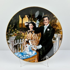 May include: Decorative plate featuring a scene with a man in a black suit and a woman in a striped dress, with a baby in a basket. The plate has a gold rim and is displayed on a clear stand. The scene is set outdoors with a white fence and greenery.