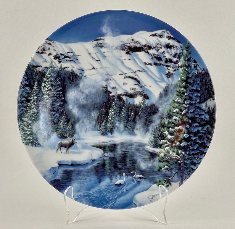 May include: A white ceramic plate with a winter scene. The plate features a snowy mountain range, a frozen lake, a lone elk, and two swans. The plate is decorated with a blue and white glaze.