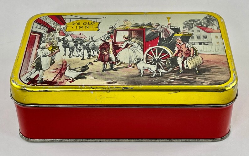 May include: A vintage tin box with a red base and a yellow lid. The lid features a colorful illustration of a horse-drawn carriage with passengers, a sign that reads "Ye Old Inn", and a group of people standing outside a building.