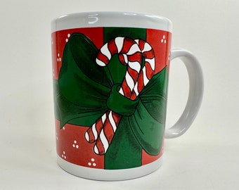 May include: White ceramic mug with a red and green Christmas-themed design. The design features a green bow with two candy canes tied together.