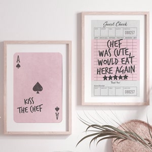Könnte beinhalten: Ein rosa gerahmter Druck mit einer schwarzen Pik-Ass-Spielkarte und dem Text "Kiss the Chef". Ein zweiter gerahmter Druck mit einem schwarz-weißen Gastcheck mit dem Text "Chef war süß, würde hier wieder essen" und fünf Sternen. Die Drucke hängen an einer weißen Wand über einer Holzbank mit einem weißen Kissen und zwei geflochtenen Körben.