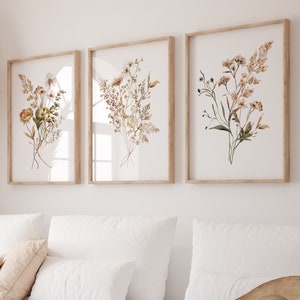Könnte beinhalten: Ein weißes Ecksofa mit drei Kissen und einem braunen geflochtenen Hut auf der Chaiselongue. Drei gerahmte botanische Drucke mit getrockneten Blumen hängen an der Wand hinter dem Sofa.