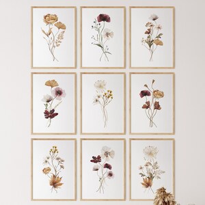 Puede incluir: Nueve impresiones de acuarela enmarcadas de diferentes arreglos florales en tonos marrones, blancos y burdeos. Las impresiones están dispuestas en una cuadrícula en una pared blanca.