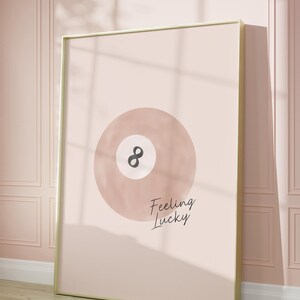 Pode incluir: Uma ilustra&ccedil;&atilde;o em aquarela rosa e branca de uma bola de bilhar com o n&uacute;mero oito nela. O texto "Feeling Lucky" est&aacute; escrito abaixo da bola.