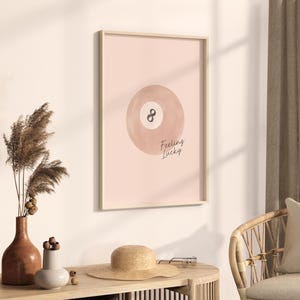 Pode incluir: Um p&ocirc;ster rosa e marrom com uma ilustra&ccedil;&atilde;o em aquarela de uma bola de bilhar com o n&uacute;mero 8 nela. O texto "Feeling Lucky" est&aacute; escrito abaixo da bola.