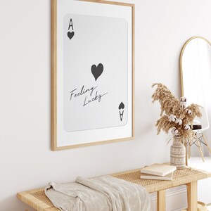 May include: A framed print of an ace of hearts playing card with the text "Feeling Lucky" written on it. The print is hanging on a white wall above a wooden bench with a woven seat and a white throw blanket.