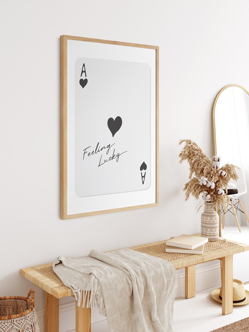 May include: A framed print of an ace of hearts playing card with the text "Feeling Lucky" written on it. The print is hanging on a white wall above a wooden bench with a woven seat and a white throw blanket.
