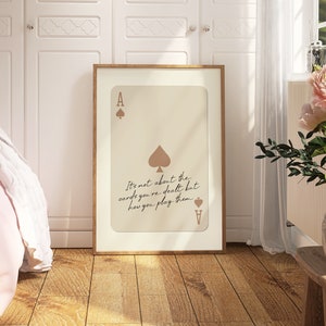 May include: A framed print with a beige background featuring an ace of spades playing card with the quote "It's not about the cards you're dealt but how you play them."