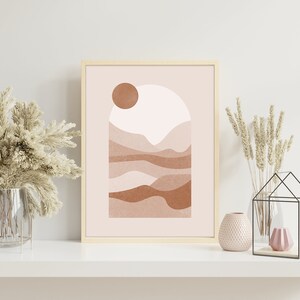 May include: A framed print with a minimalist abstract design featuring a sun, mountains, and a pink and brown color scheme. The print is displayed on a white shelf with decorative vases and plants.