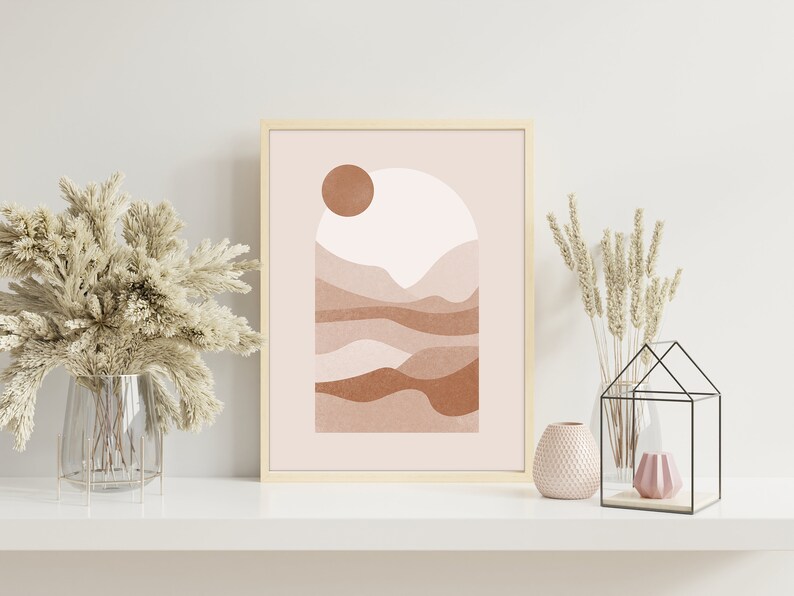 May include: A framed print with a minimalist abstract design featuring a sun, mountains, and a pink and brown color scheme. The print is displayed on a white shelf with decorative vases and plants.