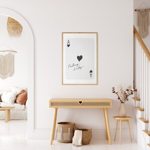 May include: A framed print of an ace of spades playing card with the text "Feeling Lucky" written on it. The print is hanging on a white wall above a wooden console table with two woven baskets and a white towel.