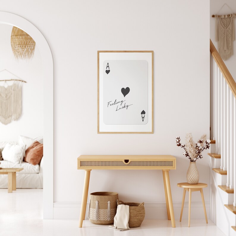 May include: A framed print of an ace of spades playing card with the text "Feeling Lucky" written on it. The print is hanging on a white wall above a wooden console table with two woven baskets and a white towel.