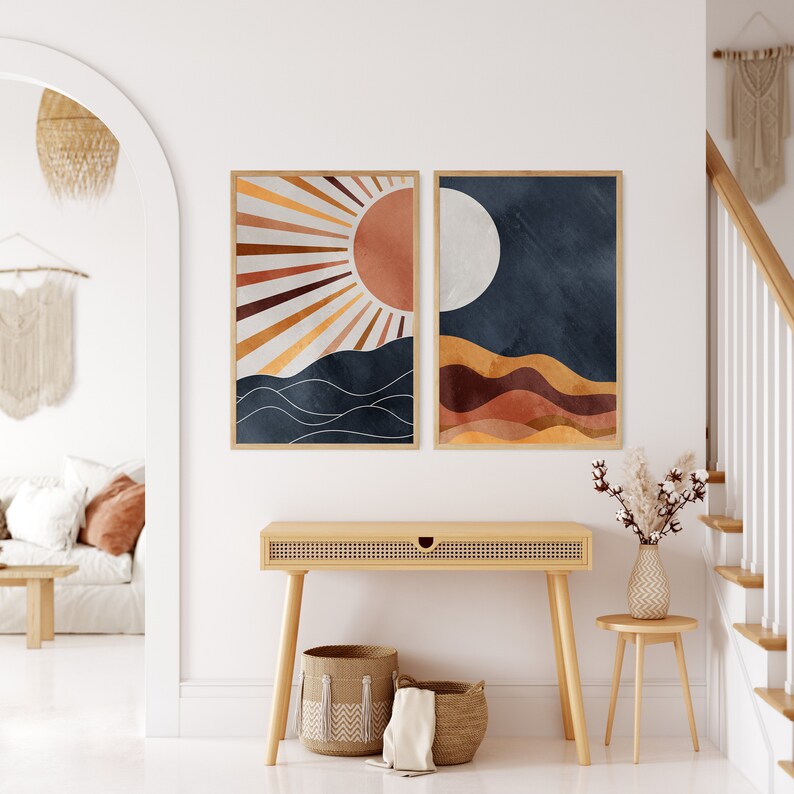 Peut inclure: Deux impressions d'art abstraites encadr&eacute;es. L'une pr&eacute;sente un motif de soleil ensoleill&eacute; dans des tons orange et marron au-dessus de vagues bleu fonc&eacute;. L'autre repr&eacute;sente une lune au-dessus de collines en couches aux tons terre. Les deux sont dans des cadres en bois clair.