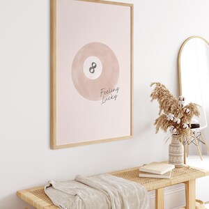 Pode incluir: Uma impress&atilde;o emoldurada com fundo rosa e uma ilustra&ccedil;&atilde;o em aquarela de uma bola de bilhar com o n&uacute;mero 8. O texto "Feeling Lucky" est&aacute; escrito em it&aacute;lico abaixo da bola.
