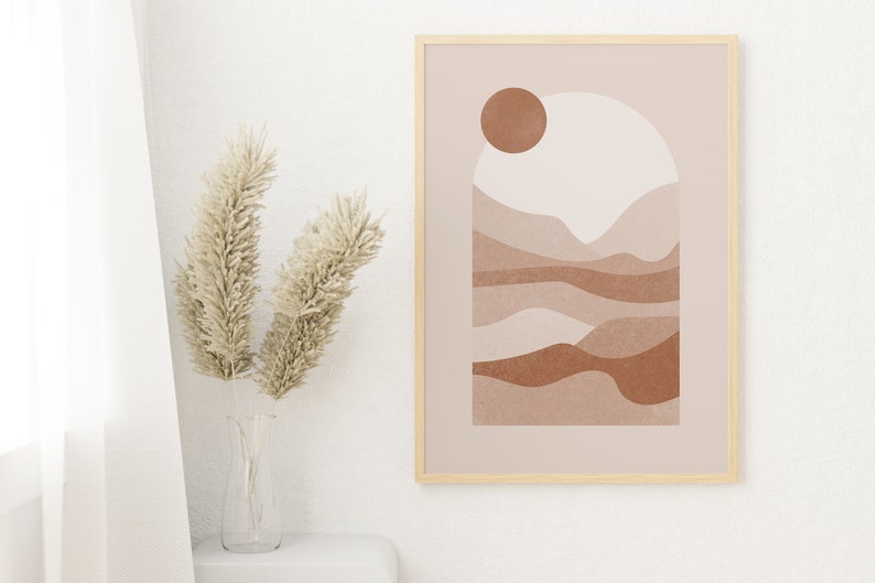 May include: A framed abstract print with a beige background and a brown sun. The print features a stylized landscape with brown and beige hills and valleys.