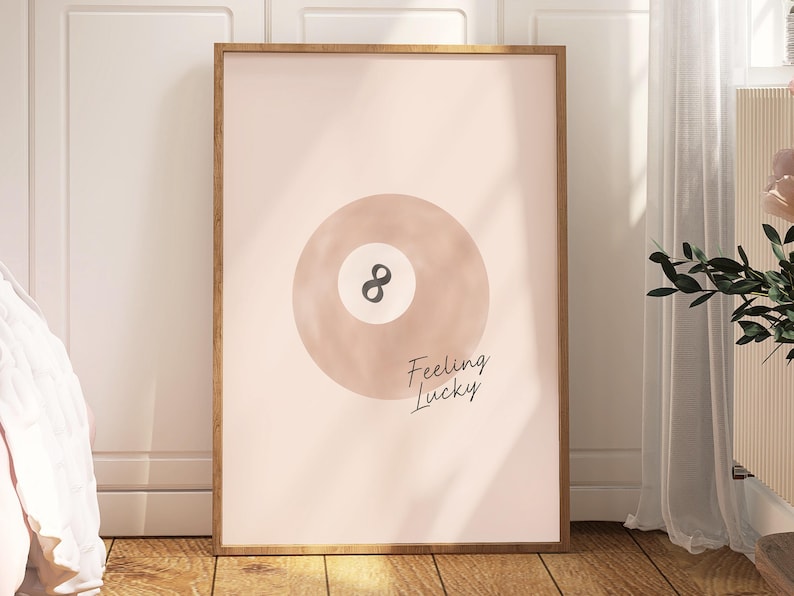 Pode incluir: Um p&ocirc;ster rosa e branco com uma ilustra&ccedil;&atilde;o em aquarela de uma bola de bilhar com o n&uacute;mero 8 nela. O texto "Feeling Lucky" est&aacute; escrito abaixo da bola.