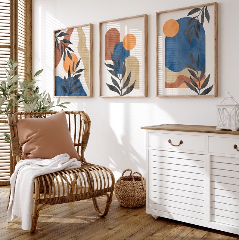 Peut inclure: Trois impressions d'art abstraites encadr&eacute;es avec des motifs &agrave; l'aquarelle dans des tons de bleu, orange et beige. Les impressions pr&eacute;sentent des &eacute;l&eacute;ments botaniques et des formes g&eacute;om&eacute;triques. Un fauteuil en osier avec un coussin et une couverture est au premier plan.