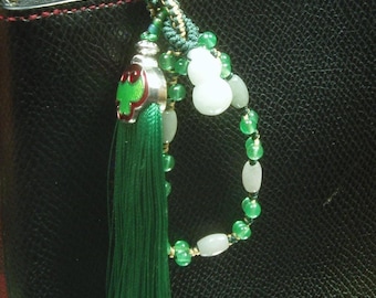 May include: A decorative charm featuring a long dark green tassel attached to a silver-toned metal cap with green enamel accents. A loop of dark green cord holds a series of small translucent green beads, opaque white beads, and a white gourd-shaped bead.