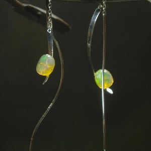 Può includere: Un paio di orecchini in argento dal design minimalista. Ogni orecchino presenta un filo d'argento curvo con una piccola pietra preziosa di opale ovale. Gli opali mostrano un mix di colori giallo, verde e bianco, creando un look sottile ed elegante.