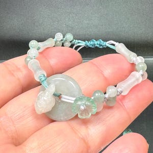 May include: A jade bracelet featuring a central, light green, circular pendant. The bracelet includes small, round beads and carved, elongated beads. A small turtle and flower are also carved into the jade. The bracelet is strung on a teal cord.