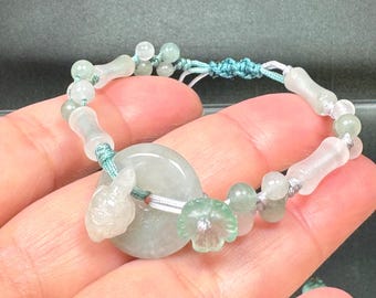 May include: A bracelet featuring a central pale green jade-like disc, a small carved stone animal, and a carved flower bead. The piece is strung on a light blue cord with alternating translucent cylindrical and spherical beads in shades of green.