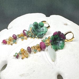 May include: A pair of gold hoop earrings featuring carved green and magenta floral gemstone charms, accented with dangling faceted ruby, emerald, and opal beads. The earrings include small clear stones on the hoops and a pale white leaf-shaped pendant.