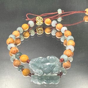 May include: A beaded bracelet featuring alternating round light brown wooden beads and translucent pale green jade beads. A central carved green jade creature charm is flanked by two gold-toned metal beads with intricate patterns on a brown cord.