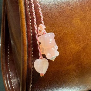以下が含まれることがあります： 薄いピンク色の半透明なクマの形をしたチャームとハート型のチャームが、細いピンク色のツイストコードにぶら下がっています。クマのチャームの上部には小さな花の彫刻が施されており、どちらも滑らかに磨かれた石で作られています。