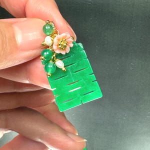 May include: A rectangular green jade pendant featuring the Chinese double happiness character. The top is adorned with a cluster of small green jade beads, white pearls, and a carved light pink flower, all connected by gold-toned metal findings.