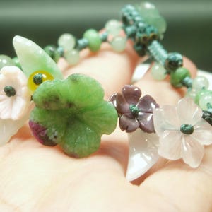 以下が含まれることがあります： 緑、ピンク、紫の色合いで彫刻された花と葉のアクセントが特徴の手作りのブレスレット。ブレスレットには、さまざまな緑の色合いの小さな丸いビーズが連なり、ターコイズのコードで繋がれています。
