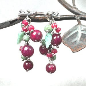 Puede incluir: Un par de pendientes colgantes con racimos de cuentas rojas oscuras, verdes y rosas. Los pendientes tienen una parte superior de metal plateado con un diseño en forma de rama. Las cuentas están dispuestas para parecerse a uvas y hojas.