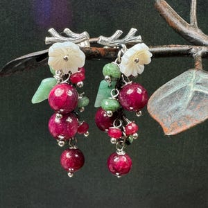 Pode incluir: Brincos pendurados com um aglomerado de pedras preciosas redondas vermelho escuro, acentuadas com pequenas pedras verdes e detalhes brancos em forma de flor. Os brincos têm detalhes em metal prateado e são projetados para se assemelhar a um cacho de uvas.
